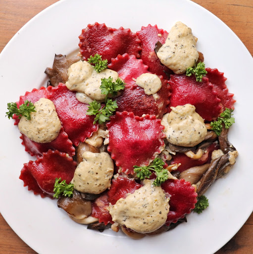 Red heart-shaped ravioli is piled high on a white plate. It is garnished with lots of NONA Alfredo, sauteed mushrooms, and garnished with fresh parsley.
