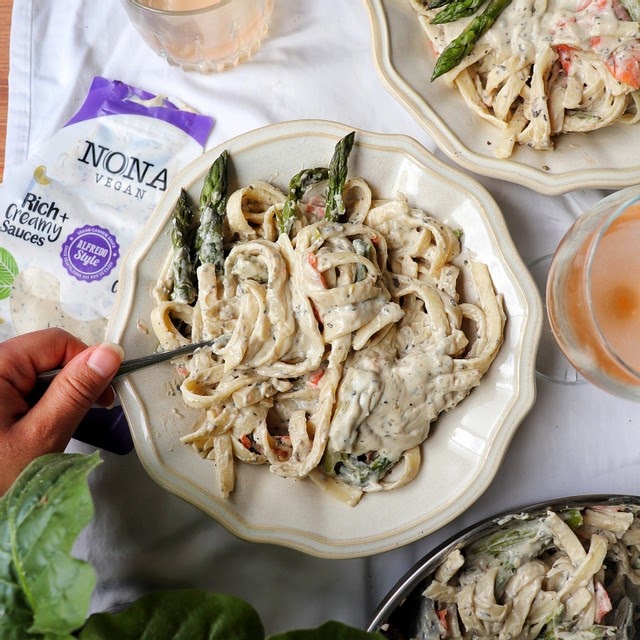 NONA Alfredo covered fettuccine noodles are heaped onto a white plate, with a side of three asparagus spears. A hand holding a fork is starting to dig-in to the deliciousness!
