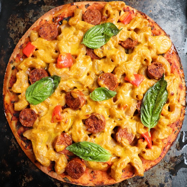 Black baking pan, Vegan pizza using a thin cauliflower crust, topped with macaroni noodles, NONA Cheesy-Style sauce, vegan sausage, and fresh basil.