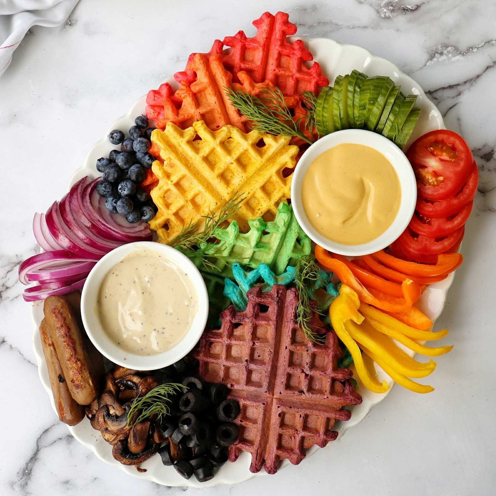 NONA's waffle board resembles a rainbow of food: red, yellow, green, blue, and purple waffles are surrounded by blueberries, vegan sausages, cucumbers, bell peppers, and blueberries. Warmed NONA sauces are nestled next to the waffles in white ramekins.