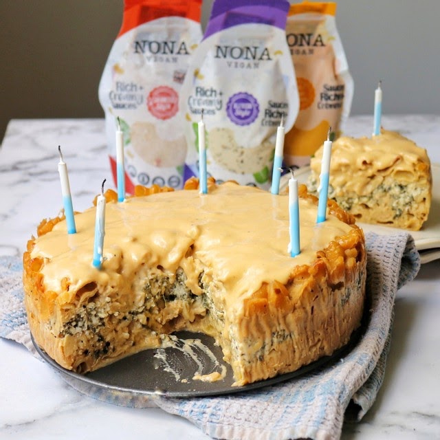 A pasta cake slathered in NONA Cheesy-Style sauce and eight blue birthday candles. In the background are NONA Carbonara, Alfredo, and Cheesy pouches.