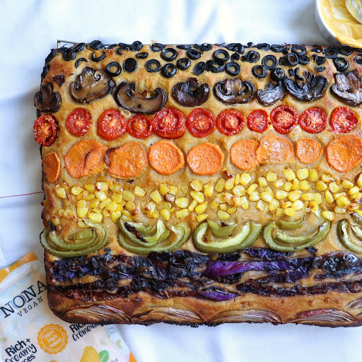 Baked focaccia line with colourful veggies to resemble the rainbow pride flag. Arranged from top to bottom: black olives, sliced mushrooms, sliced tomatoes, sweet potato coins, corn pieces, green pepper slices, strips of purple cabbage and red onion.