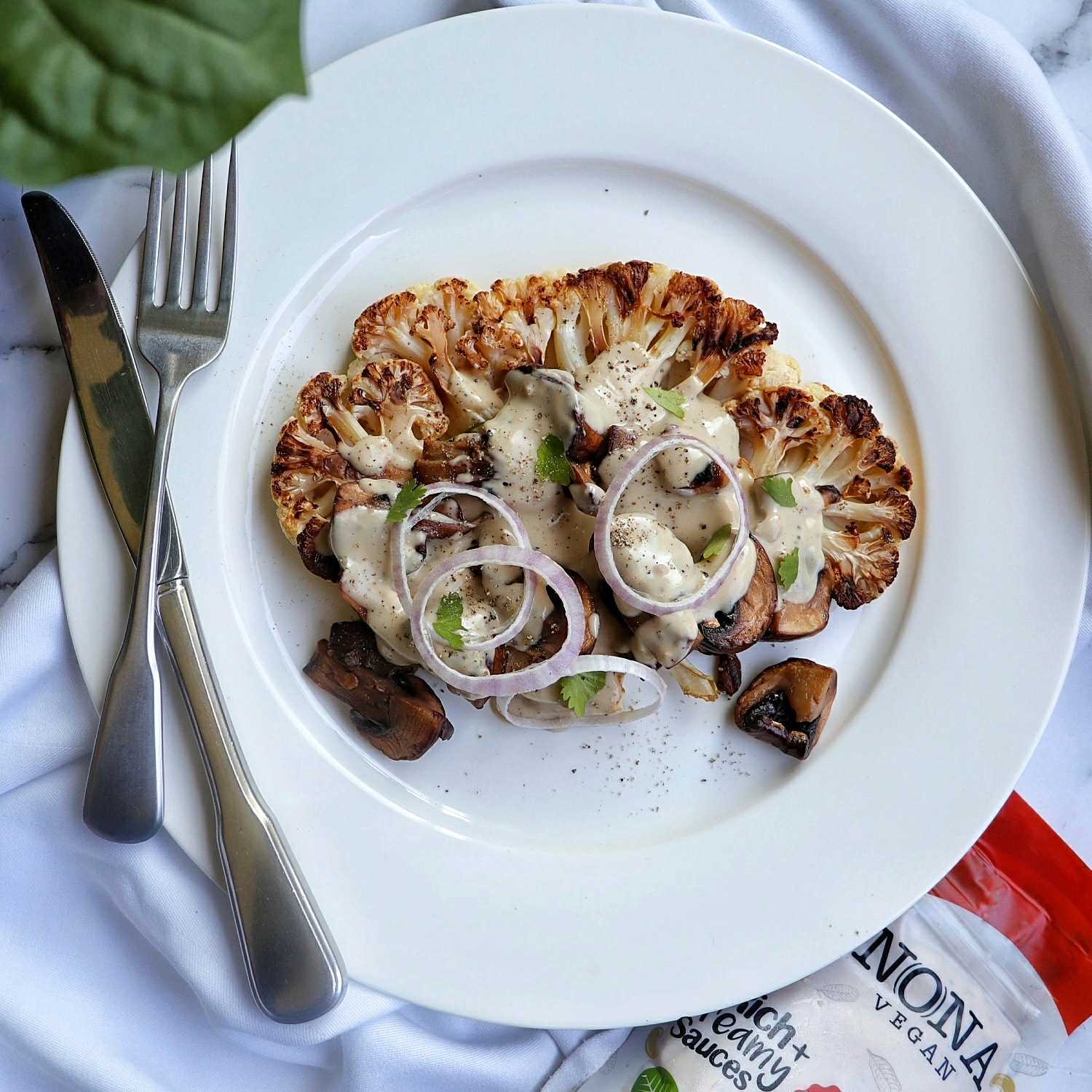 Resting on a white plate is a wide slice of cauliflower, slathered in NONA's Carbonara-style sauce, golden roasted mushrooms, and three rings of fresh red onion.