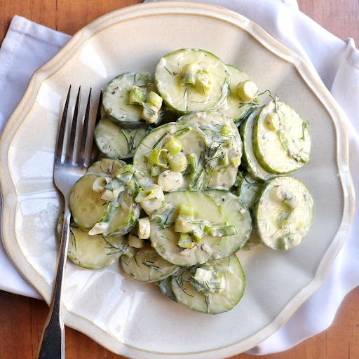 A cucumber salad, made of thinly slcied cucmbers tossed in NONA's Alfredo with dill and green onions, rests on a white scallop-edged plate. A fork is waiting to dig in!